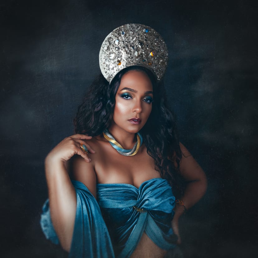 Retrato de estudio de una mujer con un tocado semicircular incrustado de piedras, gargantilla de malla dorada y azul, y un drapeado de terciopelo azul. Fondo oscuro y pictórico.